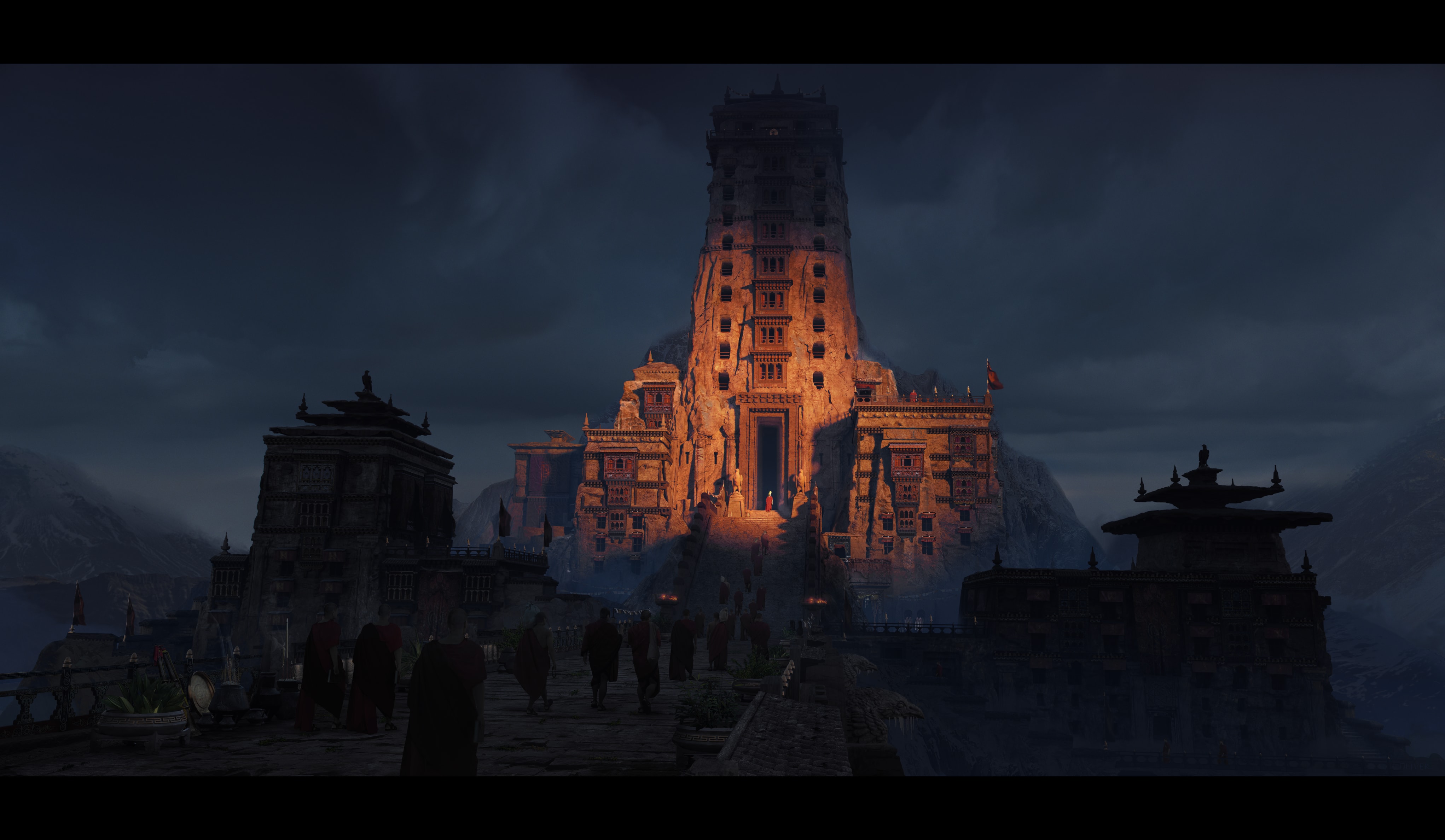 A monumental rock-cut tower temple glowing from within at dusk, as red-robed monks process toward its entrance across a stone terrace.