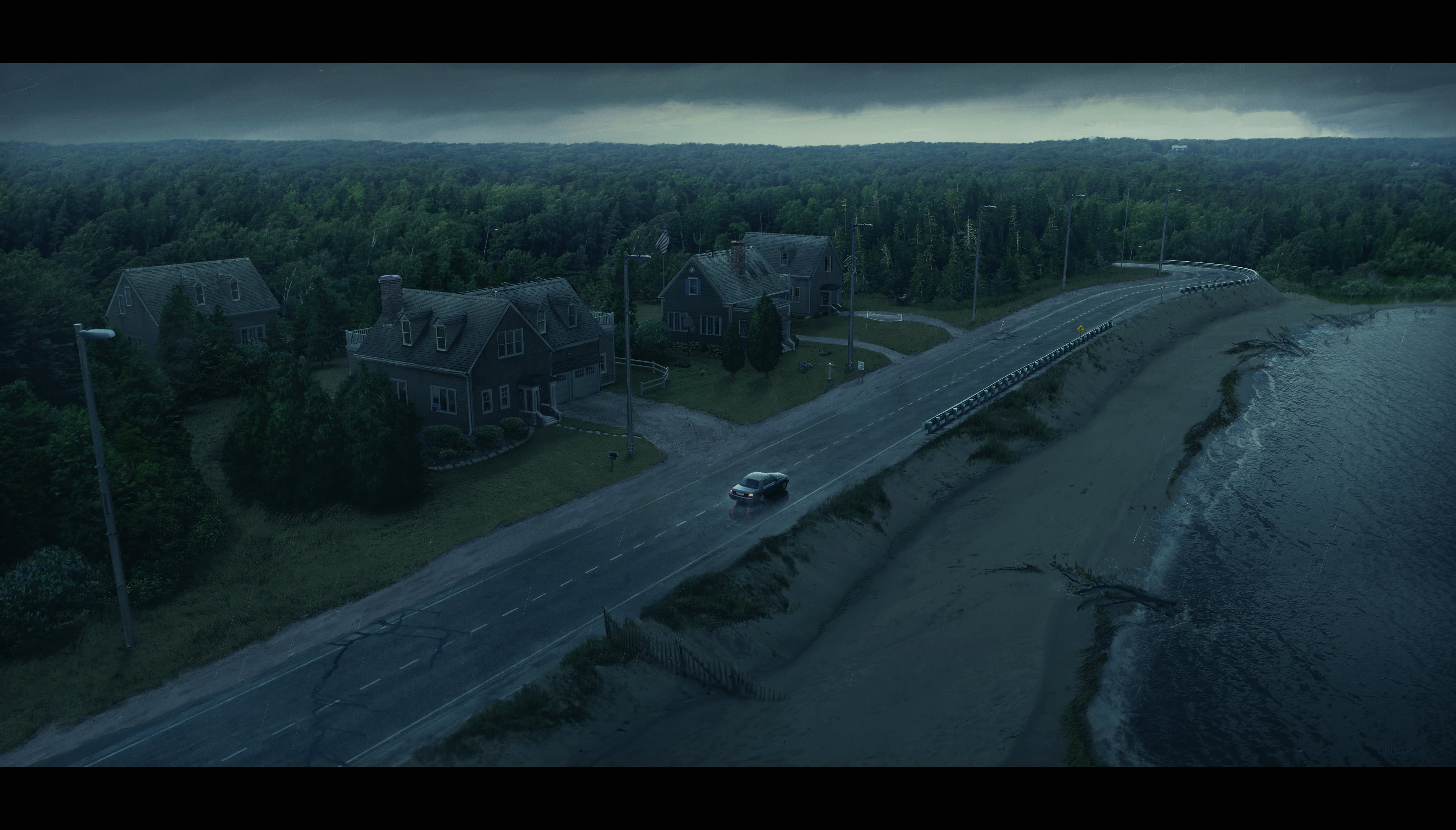 An aerial drone perspective shot of a car driving on a remote Cape Cod road. There are a row of houses to the left, and a small pond to the right. The horizon is filled with pine trees against a twilight sky with oncoming rain.